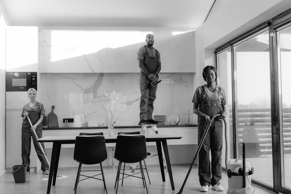 A team of three professional cleaners in a modern kitchen preparing for surface cleaning and deep cleaning tasks. The kitchen features a marble backsplash and white cabinetry, with stainless steel appliances including a microwave visible on the left. One cleaner stands on a ladder, holding a cleaning cloth, while another stands near the oven holding a mop, and the third is in front of sliding glass doors, vacuuming the wooden floor. Natural light filters through the large windows, illuminating the space, which appears spotless and well-maintained. The scene reflects hygiene and thorough cleaning practices, highlighting the importance of surface sanitisation in domestic and commercial environments, and representing services offered by Merton Cleaner as part of end-of-lease cleaning routines.