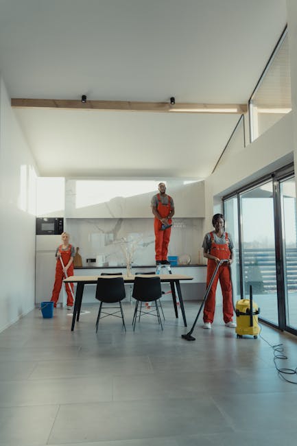 Two professional cleaners in beige uniforms stand back-to-back against a plain white wall, each holding cleaning tools. The cleaner on the left has dark hair pulled back and is holding a vacuum cleaner with a black hose and red body, suitable for surface cleaning and dust removal. The cleaner on the right has blonde hair styled in a short bob and is holding a green-handled mop, indicating floor cleaning and sanitisation. The scene emphasizes cleanliness and maintenance, reflecting a focus on domestic or commercial deep cleaning, with natural, bright lighting highlighting the pristine, dust-free surfaces. Merton Cleaner offers comprehensive cleaning services, including end-of-lease cleaning, ensuring surfaces such as floors, walls, and fixtures are thoroughly cleaned and hygienic.