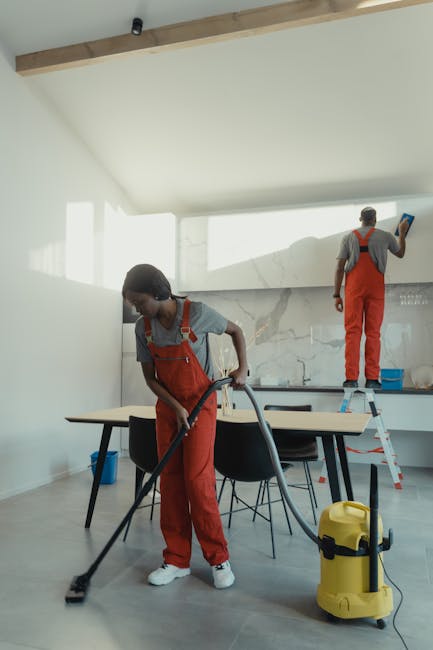 Two professional cleaners from Merton Cleaner are performing surface cleaning and sanitisation in a modern kitchen. One cleaner, a woman, is vacuuming the tiled floor with a portable vacuum cleaner, e