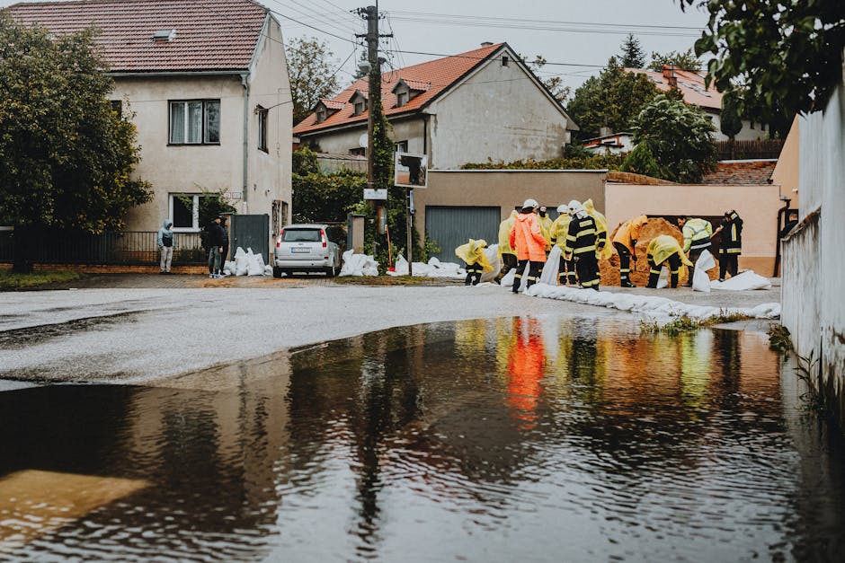 A group of workers in high-visibility jackets and protective clothing are engaged in emergency flood cleanup on a residential street, working near standing water that reflects their figures and surrounding houses. The image captures a damp pavement with a large puddle extending across the road, with some areas appearing muddy and waterlogged. Several individuals are using tools and sandbags, visible along the edge of the floodwaters, to contain or divert water flow. The background features multi-story houses with tiled roofs, some with windows and balconies, under overcast skies. Merton Cleaner’s logo and branding are not visible, but the scene emphasizes surface cleaning, flood decontamination, and water removal processes associated with emergency flood response, highlighting the need for professional sanitisation and maintenance in affected residential areas.