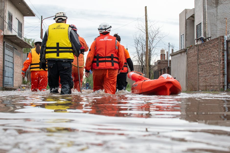 A team of emergency responders in protective helmets and high-visibility vests perform flood rescue in a residential area with flooded streets. They are walking through water that reaches their ankles, around submerged cars and partially submerged brick buildings. The responders are using a rescue floatation device and are prepared for water-based operations, with overcast skies overhead. This scene highlights the importance of professional flood response and surface water management, similar to the emergency flood cleanup service options offered by Merton Cleaner in Merton. The image emphasizes safety, water removal, and the importance of rapid response in flood-affected areas.