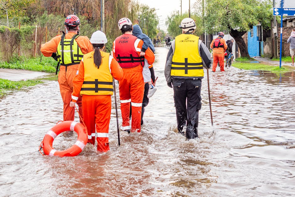 A group of workers in high-visibility jackets and protective clothing are engaged in emergency flood cleanup on a residential street, working near standing water that reflects their figures and surrounding houses. The image captures a damp pavement with a large puddle extending across the road, with some areas appearing muddy and waterlogged. Several individuals are using tools and sandbags, visible along the edge of the floodwaters, to contain or divert water flow. The background features multi-story houses with tiled roofs, some with windows and balconies, under overcast skies. Merton Cleaner’s logo and branding are not visible, but the scene emphasizes surface cleaning, flood decontamination, and water removal processes associated with emergency flood response, highlighting the need for professional sanitisation and maintenance in affected residential areas.