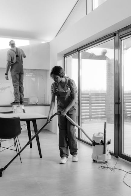 Two professional cleaners, both wearing grey shirts and bright orange overalls, perform surface cleaning in a modern living room. The cleaner on the left is vacuuming the polished, light grey tiled fl
