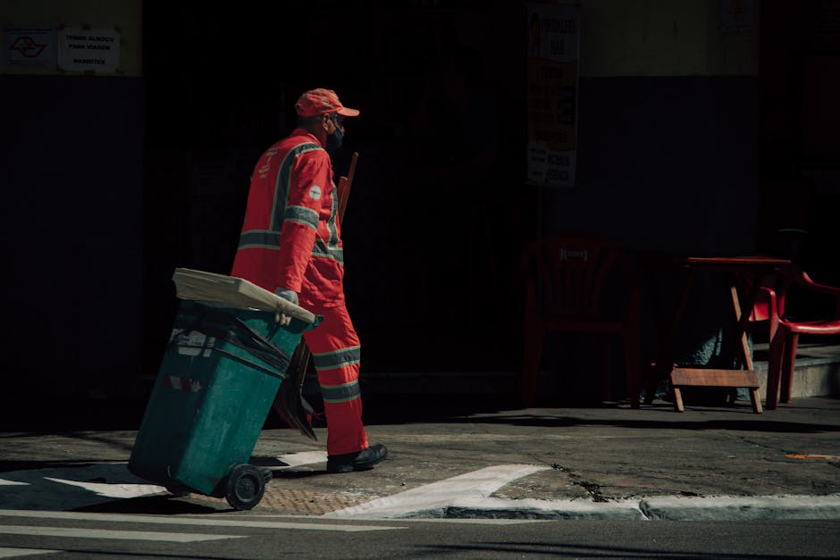 A street cleaning worker dressed in a red uniform with reflective strips is walking along a pavement, pushing a large teal wheeled trash bin. The worker is wearing a cap and a mask, and is illuminated by natural sunlight, casting shadows on the ground. In the background, there are some red plastic chairs and a table outdoors, with a dark building facade behind them. The scene appears clean and orderly, highlighting street maintenance and waste management activities that are essential for urban hygiene; this description aligns with surface cleaning and sanitation practices discussed by Merton Cleaner in relation to commercial waste rules.