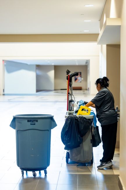 A mobile cleaning worker dressed in an orange uniform holding two large blue trash bags, standing on a smooth gray floor against a plain white wall, with a black and orange spray bottle with a connected hose or nozzle in the foreground. The scene is well-lit, showcasing the clean and organized environment typical of commercial waste management and surface cleaning, emphasizing hygienic disposal practices in line with Merton Council's commercial waste rules. Merton Cleaner provides professional cleaning services focusing on waste removal, deep cleaning, and sanitisation to maintain hygiene standards in both residential and commercial settings.