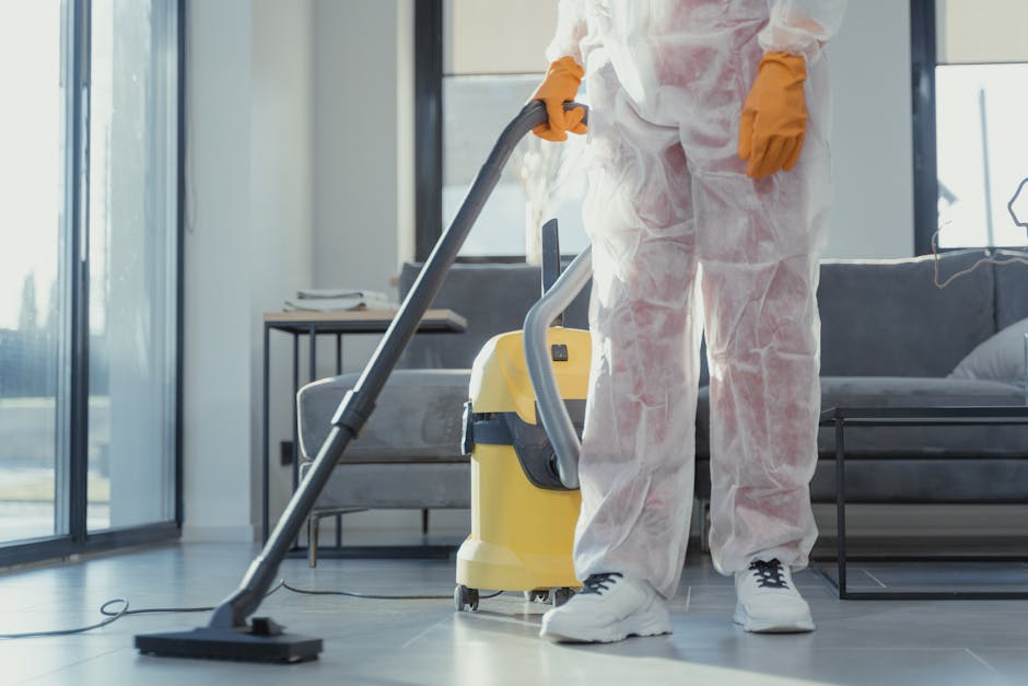 A professional cleaner dressed in white protective coveralls and orange gloves is operating a yellow and gray vacuum cleaner on a dark tiled floor in a modern living room. The room features large floor-to-ceiling windows allowing natural light to fill the space, with a gray sofa and a small side table visible in the background. The cleaner is focused on surface cleaning, ensuring the floors are free of dust and debris. The setting highlights a clean, tidy environment with a focus on deep cleaning and sanitisation, reflecting the services offered by Merton Cleaner for domestic cleaning and waste removal in Mitcham. The scene emphasizes detailed surface cleaning in a contemporary home setting.