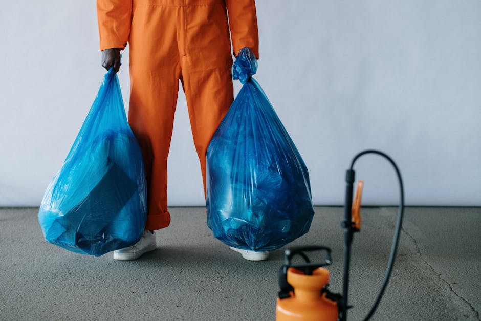 A young woman with a cheerful expression is standing against a plain white background, dressed casually in a plaid shirt, gray t-shirt, and light blue jeans. She is wearing pink rubber gloves and holding a white plastic bottle with a green recycling symbol, suggesting eco-friendly cleaning. The scene emphasizes surface cleaning and sanitisation, aligned with domestic cleaning services offered by Merton Cleaner, promoting hygiene and sustainable practices in residential or commercial cleaning contexts.