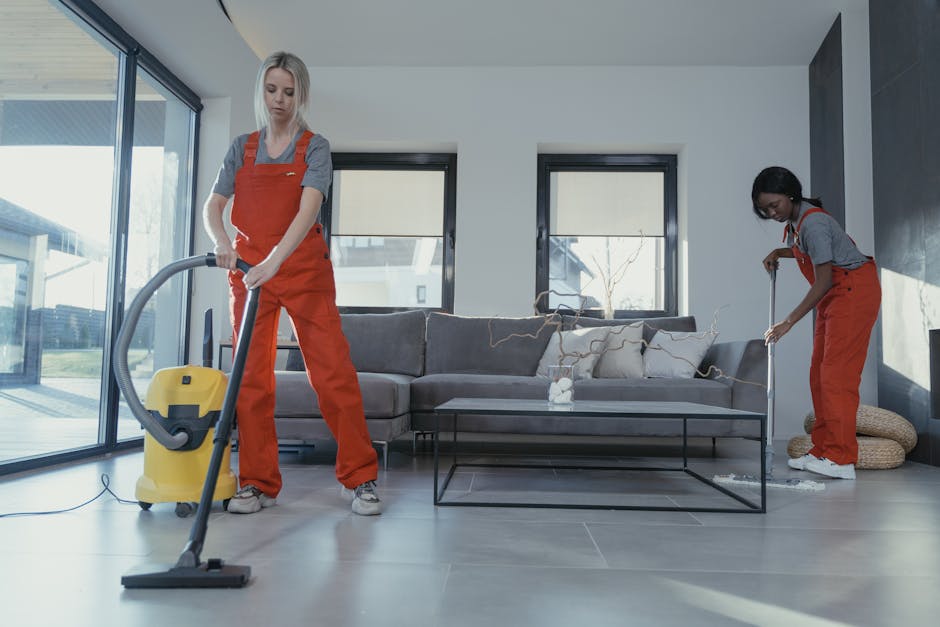 Two professional cleaners wearing orange overalls and gray shirts performing a deep cleaning task in a modern living room. One cleaner is using a yellow vacuum cleaner on the light gray tiled floor, while the other is using a mop to clean the area around a gray sofa with beige and white cushions. The room features large windows with natural light, a black metal coffee table with a glass top, and a dark accent wall. The surfaces appear dust-free and shiny, indicating thorough surface cleaning and maintenance, reflecting a high standard of hygiene in domestic cleaning. The scene emphasizes detailed cleaning processes in residential spaces, consistent with the services offered by Merton Cleaner.