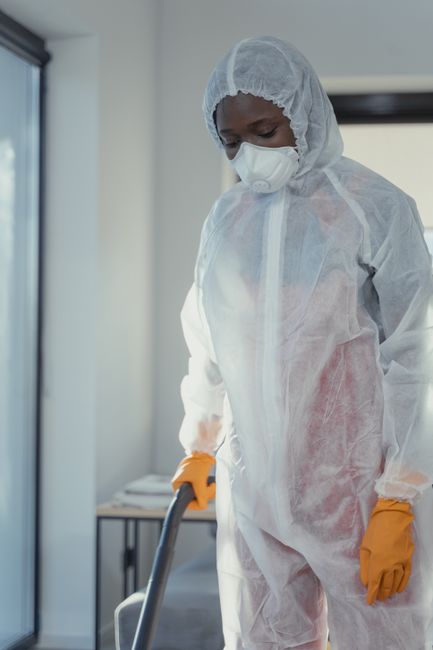 A professional cleaner dressed in a white protective suit with a hood, a face mask, and orange gloves is using a vacuum cleaner on a floor in an indoor setting. The surface appears to be a smooth, hard material, possibly wood or tile, with some dust or debris visible. The cleaning area is well-lit with natural light coming through a window or glass door in the background, and the room has minimal furniture, indicating a deep cleaning or sanitisation process. This scene demonstrates surface cleaning and hygiene practices typical of residential or commercial cleaning services, as featured by Merton Cleaner, aligned with odour removal for damp flats near Wimbledon Common.