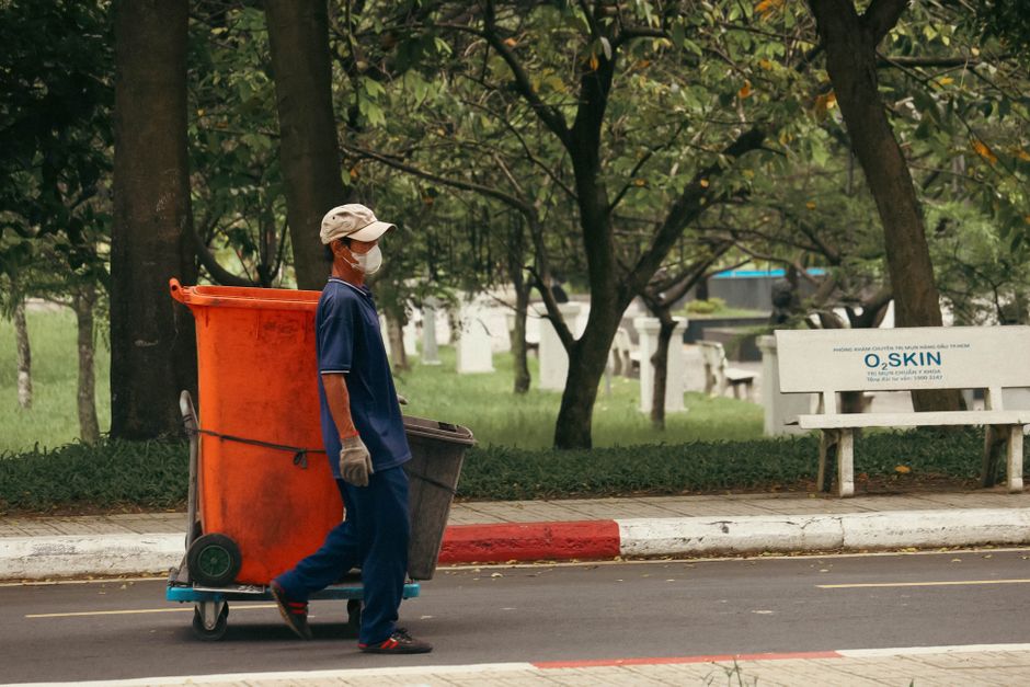 A street cleaner in a blue uniform, wearing a face mask and gloves, is pushing a trolley with a large orange waste bin and a smaller black bin along a sidewalk. The scene is set in a park-like area with green grass, trees, and a white bench with 'O2 SKIN' signage in the background. The ground surface is paved, and the street has a white and red painted curb, suggesting an urban residential environment. The image emphasizes routine urban cleaning and sanitation efforts, highlighting the importance of hygiene in maintaining clean public spaces, in line with the cleaning services promoted by Merton Cleaner.