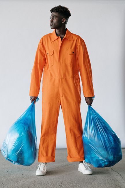 A young man with dark curly hair and medium skin tone standing on a plain light gray background, holding two large blue garbage bags filled with waste, one in each hand. He is dressed in an orange protective jumpsuit and white sneakers. The scene suggests waste collection or disposal activity, highlighting cleanliness and waste management practices relevant to domestic or commercial cleaning services. This image illustrates the importance of proper waste handling as part of professional cleaning or sanitation routines offered by Merton Cleaner, in line with advice on skip permits, fines, and disposal rules in Merton.