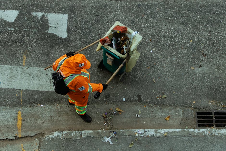 A street cleaner in a blue uniform, wearing a face mask and gloves, is pushing a trolley with a large orange waste bin and a smaller black bin along a sidewalk. The scene is set in a park-like area with green grass, trees, and a white bench with 'O2 SKIN' signage in the background. The ground surface is paved, and the street has a white and red painted curb, suggesting an urban residential environment. The image emphasizes routine urban cleaning and sanitation efforts, highlighting the importance of hygiene in maintaining clean public spaces, in line with the cleaning services promoted by Merton Cleaner.