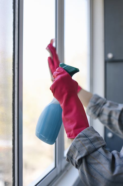Close-up of a person wearing pink rubber gloves and a plaid shirt, using a spray bottle and a cloth for surface cleaning on a large glass window in a residential or communal building. The window has a clean, shiny glass pane, allowing natural light to illuminate the area. The background outside the window is blurred, showing an outdoor scene. This image illustrates standard glass and window cleaning practices, emphasizing hygiene and maintenance essential for communal block environments, as part of a domestic or deep cleaning process by Merton Cleaner, a professional cleaning service.