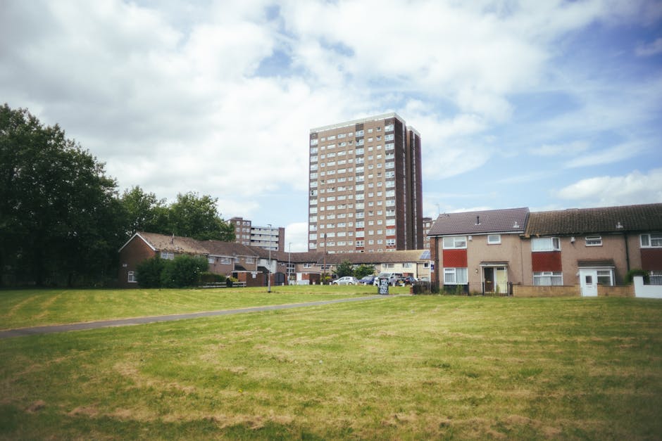 A wide view of a residential communal area with a well-maintained grassy lawn, bordered by small brick houses with tiled roofs on one side and a tall, multi-storey apartment building in the background. The scene is illuminated by natural daylight with partly cloudy skies, highlighting the clean and tidy appearance of the outdoor space. The area appears to be clear of debris or litter, reflecting effective surface cleaning and maintenance by Merton Cleaner, who specialise in sanitation and deep cleaning services for communal blocks within West Barnes Estate.