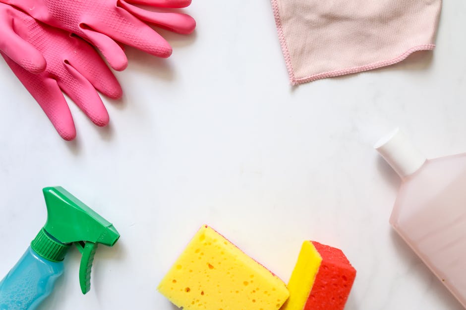 A flat lay of cleaning tools and supplies arranged on a white surface, including a pair of pink rubber gloves, a blue spray bottle with a green nozzle, a pink cloth, two sponges (one yellow and one red), and a pink plastic bottle. The items are positioned with the gloves near the top left corner, the cloth at the top right, and the cleaning solutions and sponges towards the bottom, illustrating the materials used for surface cleaning and sanitation in domestic cleaning tasks. The setup emphasizes preparation for thorough cleaning, as promoted by Merton Cleaner for residential spaces such as Wimbledon Broadway flats.